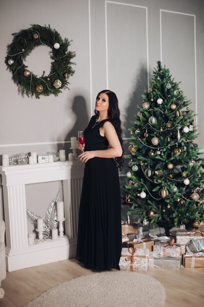 Hübsche junge kaukasische Frau mit langen dunklen Haaren im langen schwarzen Kleid steht nahe dem Weihnachtsbaum vor dem Abendessen