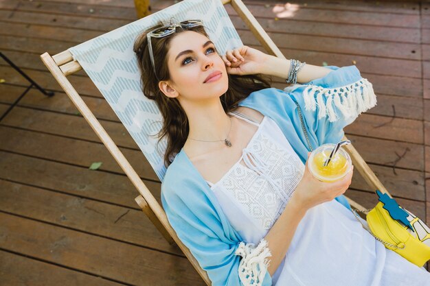 Hübsche junge lächelnde stilvolle Frau, die im Sommer-Outfit im Liegestuhl sitzt und weißes Kleid, blauen Umhang, Sonnenbrille, Geldbörse trägt, frischen Saft trinkt und sich entspannt