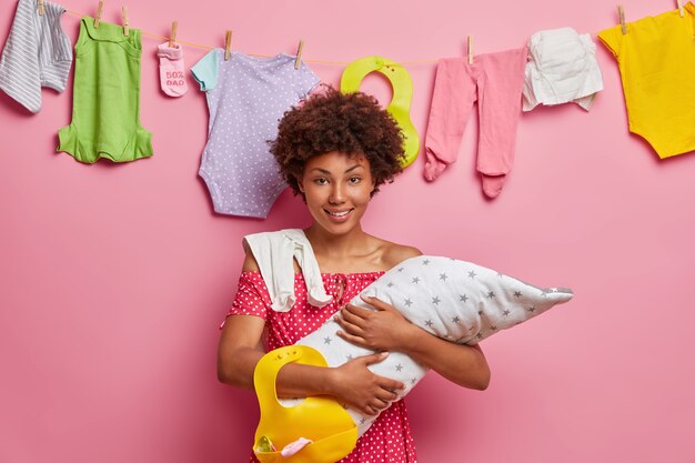 Hübsche junge Mutter mit Afro-Haaren, hält Neugeborenes in Decke gewickelt, Gummilätzchen zum Füttern des Säuglings drückt Liebe und Pflege steht