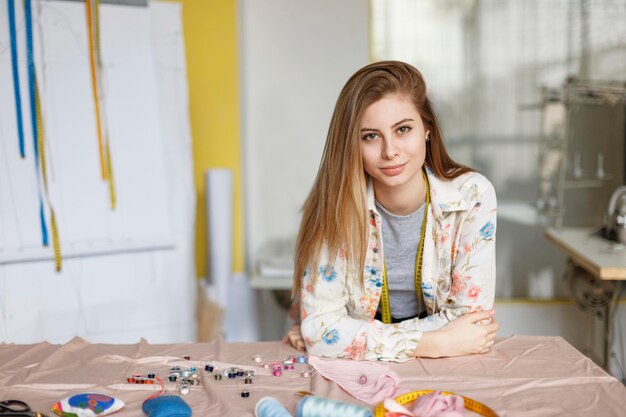 Hübsche junge Näherin im Hemd, die sich auf den Tisch lehnt und in der modernen Nähwerkstatt verträumt in die Kamera schaut