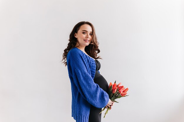 Hübsche junge schwangere Frau hält Tulpen. Brunette Mädchen in der blauen Strickjacke und im schwarzen Kleid wirft mit Blumenstrauß auf lokalisiertem Hintergrund auf.