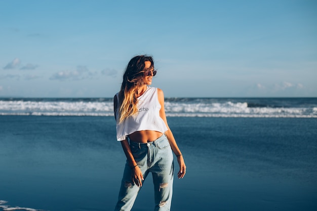 Hübsche kaukasische passende frau im weißen oberteil und in den jeans auf reflektierendem strand durch ozean bei sonnenuntergangslicht