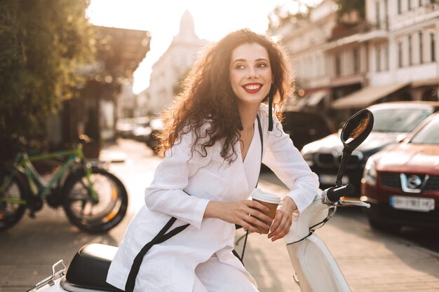 Hübsche lächelnde Dame mit dunklem, lockigem Haar in weißem Kostüm, die auf einem weißen Moped mit einer Tasse Kaffee zum Mitnehmen sitzt und glücklich mit schönem Blick auf die Stadt im Hintergrund zur Seite schaut