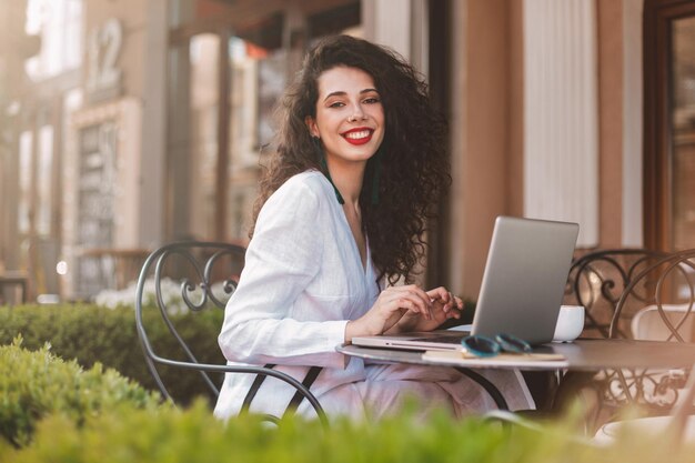 Hübsche lächelnde dame mit dunklem lockigem haar in weißem kostüm, die mit laptop am tisch sitzt und glücklich in die kamera schaut, während sie zeit im café auf der straße verbringt