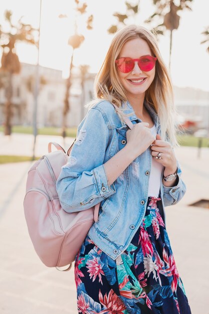 Hübsche lächelnde Frau, die in der Stadtstraße im stilvollen bedruckten Rock und in der übergroßen Jeansjacke geht, die rosa Sonnenbrille trägt, Lederrucksack hält, Sommerstil-Trend