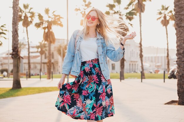 Hübsche lächelnde Frau, die in der Stadtstraße im stilvollen bedruckten Rock und in der übergroßen Jeansjacke trägt, die rosa Sonnenbrille, Sommerarttrend trägt