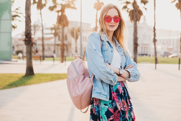 Hübsche lächelnde Frau, die in der Stadtstraße im stilvollen bedruckten Rock und in der übergroßen Jeansjacke trägt, die rosa Sonnenbrille, Sommerarttrend trägt