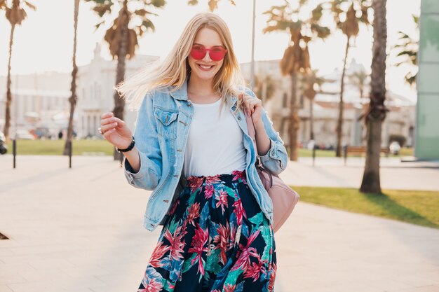 Hübsche lächelnde Frau, die in einem stilvollen bedruckten Rock und einer übergroßen Jeansjacke mit rosa Sonnenbrille in der Stadtstraße spaziert