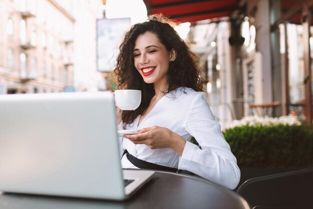 Hübsche lächelnde Frau mit dunklem lockigem Haar in weißem Kostüm, die mit einer Tasse Kaffee in den Händen am Tisch sitzt und glücklich in den Laptop schaut, während sie Zeit im Café auf der Straße verbringt