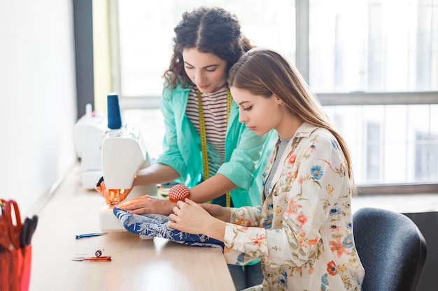 Hübsche Näherin unterrichtet Mädchen, die in Nähkursen mit Nähmaschine arbeiten, mit Fenster im Hintergrund in moderner Nähwerkstatt