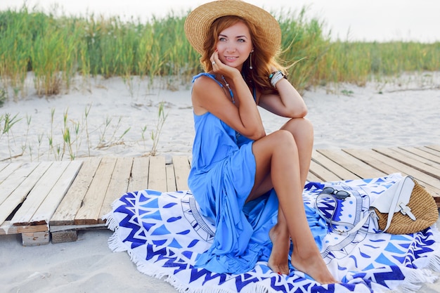 Hübsche schlanke Frau mit langen roten Haaren im Strohhut, die erstaunliche Urlaubszeit am Strand verbringen. Blaues Kleid tragen. Sitzen auf stilvollem Cover.