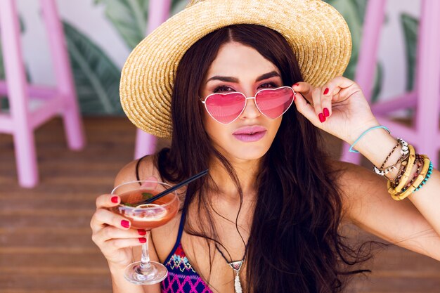 Hübsche Strandfrau in der hellen Farbe Badebekleidung, rosa Herzsonnenbrille und Strohhut, die Sommerzeit genießen
