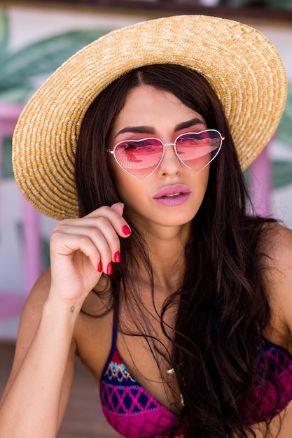 Hübsche Strandfrau in heller Badebekleidung, rosafarbener Herz-Sonnenbrille und Strohhut, die den Sommer genießen