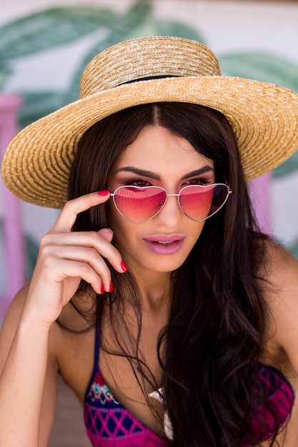 Hübsche Strandfrau in heller Badebekleidung, rosafarbener Herzsonnenbrille und Strohhut, die den Sommer genießt.