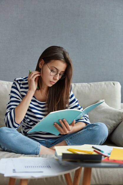 Hübsche Studentin mit dunklem Haar im Lehrbuch, sitzt gekreuzte Beine auf dem Sofa