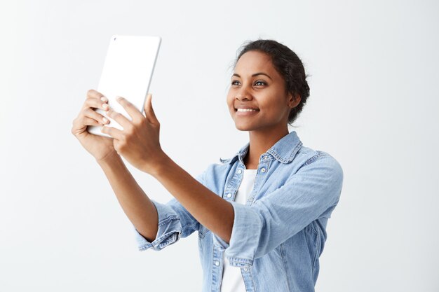 Hübsche und fröhliche afroamerikanische junge Frau im blauen Hemd über weißem T-Shirt, das für selfie aufwirft und lächelt, Tablette auf weißer Wand hält Menschen, Emotionen und moderne Technologien