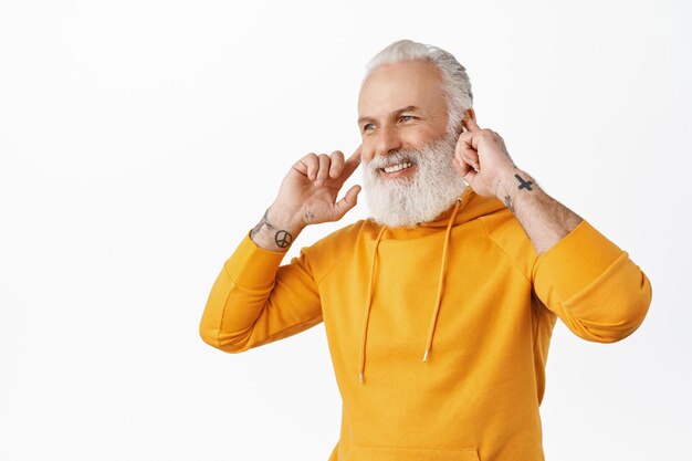 Hübscher älterer Mann hört Musik auf drahtlosen Kopfhörern und lächelt und schaut beiseite auf das Logo. Hipster alter Mann, der perfekten Klang von Ohrhörern genießt, weißer Hintergrund.