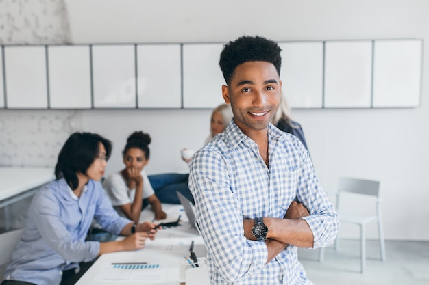 Hübscher afrikanischer Mann, der auf Tisch im Büro sitzt, während seine Untergebenen an neuer Verkaufsstrategie arbeiten. Innenporträt von Geschäftsleuten internationaler Unternehmen, die während des Arbeitsprozesses posieren.