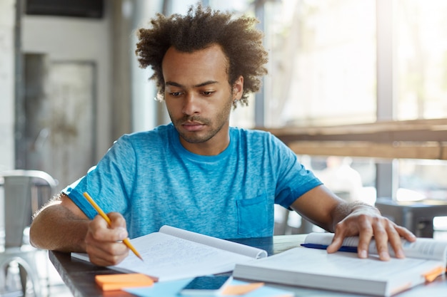 Hübscher afroamerikanischer Doktorand mit lockiger Frisur, der am Schreibtisch mit Buch und Heft sitzt, Informationen und Notizen studiert, sich auf Prüfung oder Test vorbereitet, fokussiertes und konzentriertes Aussehen hat