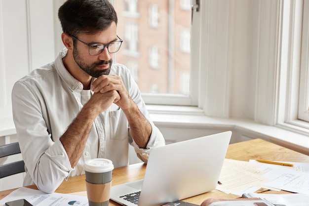 Hübscher Angestellter hat ernsthaften konzentrierten Blick auf Laptop-Computer, trägt transparente Brille und weißes Hemd, arbeitet mit Laptop-Computer,