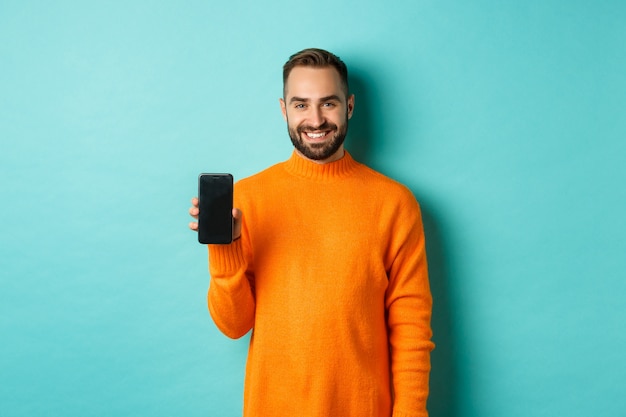 Hübscher bärtiger Kerl im orangefarbenen Pullover, zeigt Smartphonebildschirm und lächelt, zeigt Promo online, türkisfarbene Wand.
