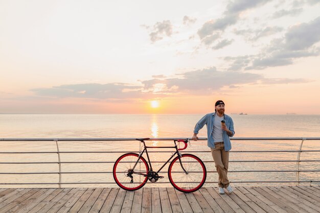 Hübscher bärtiger Mann, der mit dem Fahrrad im Morgensonnenaufgang durch das Meer reist, das Kaffee trinkt, gesunder Reisender des aktiven Lebensstils