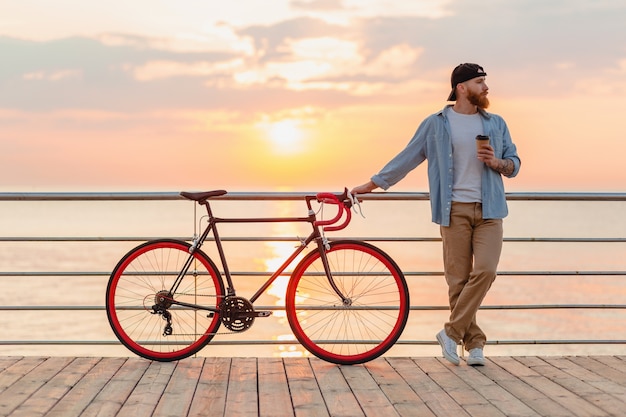 Hübscher bärtiger Mann, der mit dem Fahrrad im Morgensonnenaufgang durch das Meer reist, das Kaffee trinkt, gesunder Reisender des aktiven Lebensstils