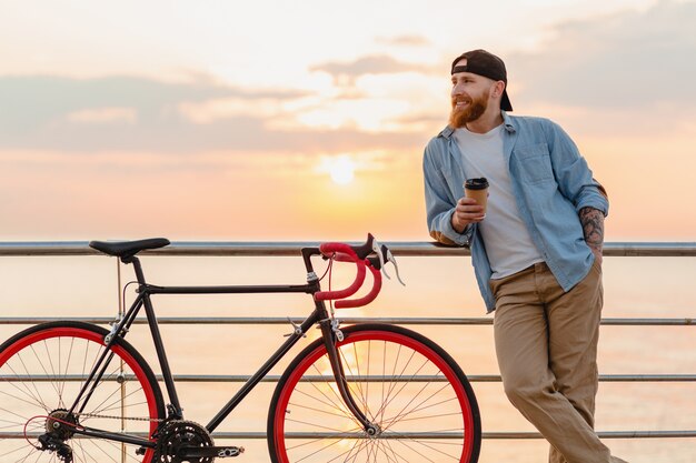 Hübscher bärtiger Mann, der mit dem Fahrrad im Morgensonnenaufgang durch das Meer reist, das Kaffee trinkt, gesunder Reisender des aktiven Lebensstils