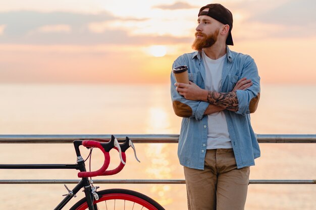 Hübscher bärtiger Mann, der mit dem Fahrrad im Morgensonnenaufgang durch das Meer reist, das Kaffee trinkt, gesunder Reisender des aktiven Lebensstils