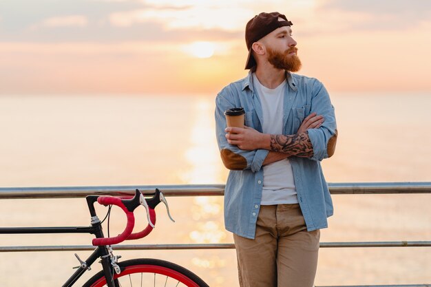 Hübscher bärtiger Mann, der mit dem Fahrrad im Morgensonnenaufgang durch das Meer reist, das Kaffee trinkt, gesunder Reisender des aktiven Lebensstils