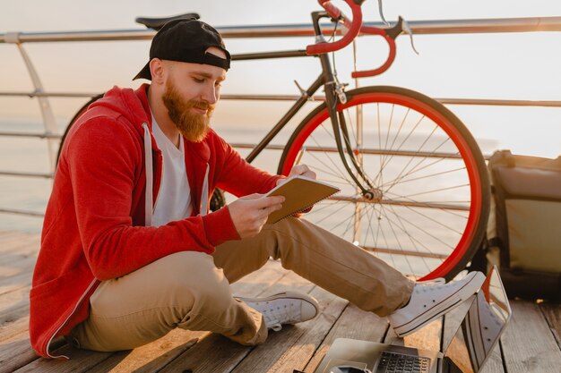 Hübscher bärtiger Mann des Hipster-Stils, der Online-Freiberufler auf Laptop mit Rucksack und Fahrrad im Morgensonnenaufgang durch den gesunden aktiven Lebensstilreisenden-Rucksacktouristen des Meeres arbeitet