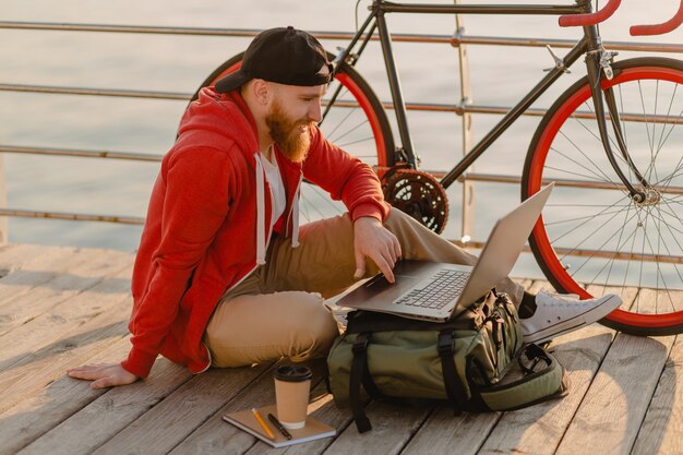 Hübscher bärtiger Mann des Hipster-Stils, der Online-Freiberufler auf Laptop mit Rucksack und Fahrrad im Morgensonnenaufgang durch den gesunden aktiven Lebensstilreisenden-Rucksacktouristen des Meeres arbeitet