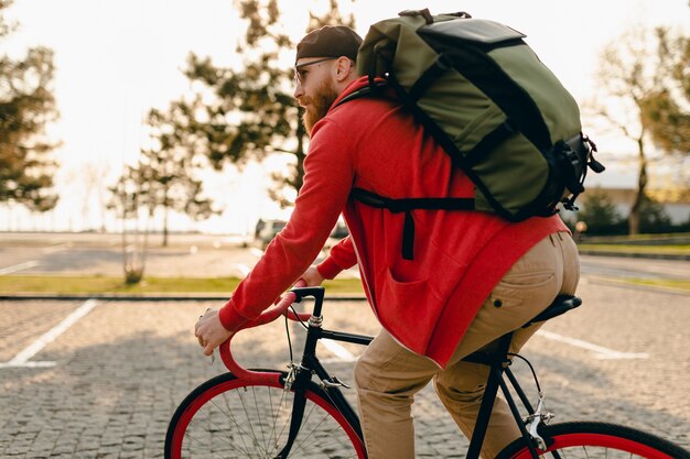 Hübscher bärtiger Mann des Hipster-Stils im roten Kapuzenpulli und in der Sonnenbrille, die allein mit Rucksack auf Fahrradreisenden Rucksacktouristen des gesunden aktiven Lebensstils reiten