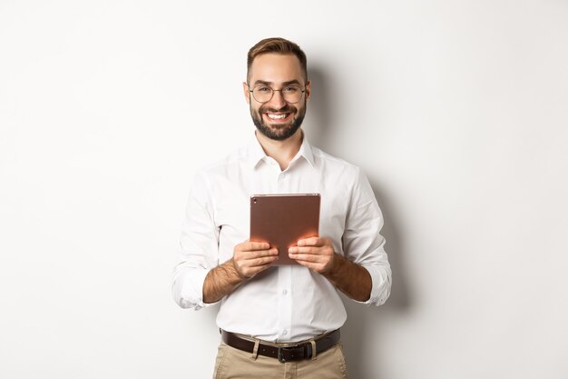 Hübscher bärtiger Mann in den Gläsern unter Verwendung der digitalen Tablette, lächelnd zufrieden, stehend