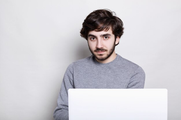 Hübscher bärtiger Mann mit sylisch dunklem Haar, der lässigen grauen Pullover trägt, der E-Mail auf Laptop überprüft