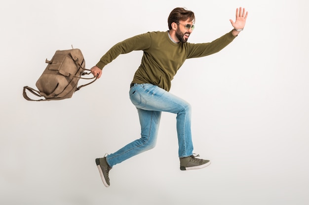 Hübscher bärtiger stilvoller Mann, der isoliert läuft, gekleidet in Sweatshirt mit Reisetasche, Jeans und Sonnenbrille tragend, verrückter Reisender auf Eile