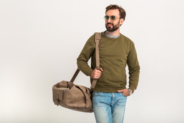 Hübscher bärtiger stilvoller Mann, der lokal gekleidet im Sweatshirt mit Reisetasche aufwirft, Jeans und Sonnenbrille tragend