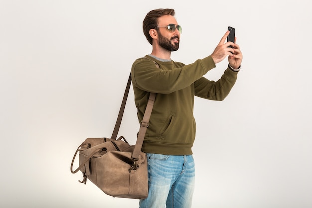 Hübscher bärtiger stilvoller Mann im Sweatshirt mit Reisetasche, tragen Jeans und Sonnenbrille lokalisiert Selfie-Foto am Telefon