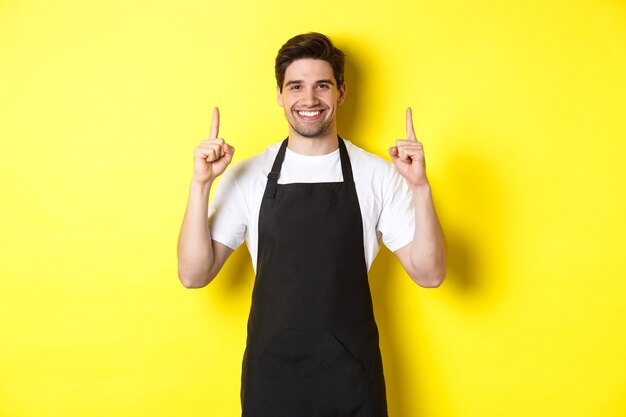 Hübscher Barista, der Werbung zeigt, Coffeeshop-Promo-Angebot, Finger nach oben zeigend, vor gelbem Hintergrund stehend.