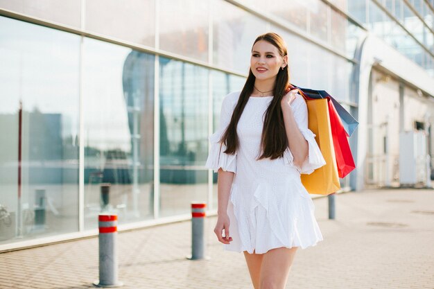 Hübscher Brunette mit dem langen Haar gehend mit Einkaufstaschen vor einem modernen Gebäude