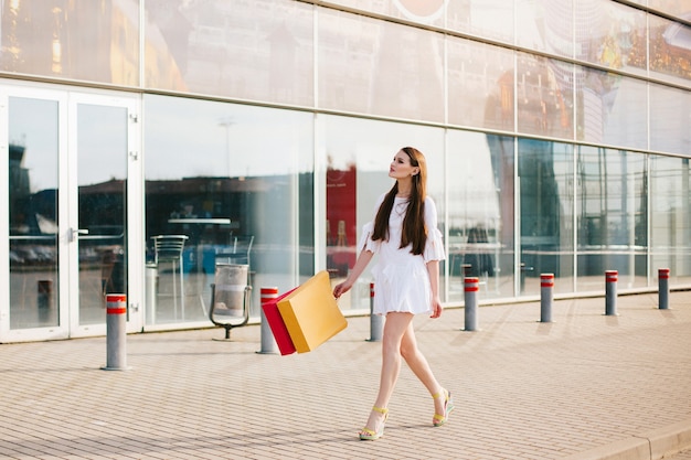 Hübscher Brunette mit dem langen Haar gehend mit Einkaufstaschen vor einem modernen Glasgebäude