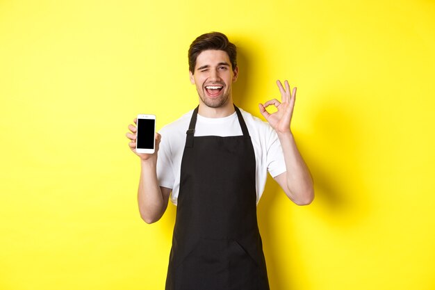 Hübscher Coffeeshop-Arbeiter, der OK-Zeichen und Smartphone-Bildschirm zeigt, Anwendung empfehlend, über gelbem Hintergrund stehend.