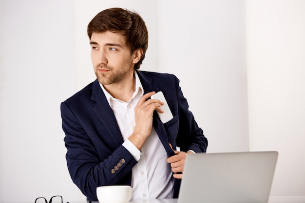 Hübscher erfolgreicher Geschäftsmann sitzen Schreibtisch, trinken Kaffee und checken Post im Laptop, stecken Handy in Jackentasche