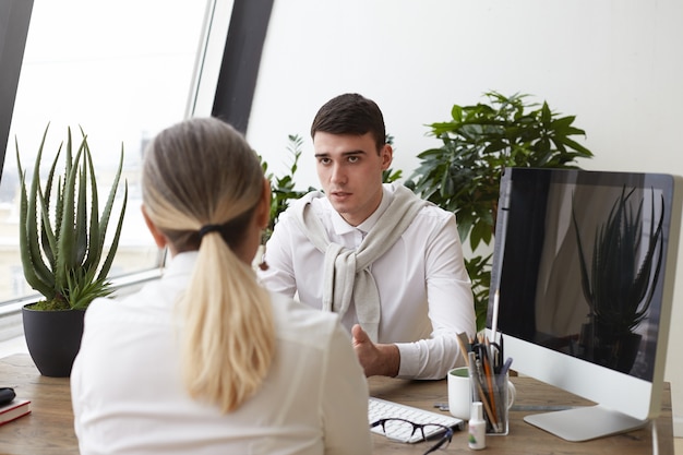 Kostenloses Foto hübscher ernsthafter junger mann hr-spezialist, der unerkennbare frau mit pferdeschwanz während des vorstellungsgesprächs interviewt und fragen über ihre erfahrungen und fähigkeiten stellt. beschäftigung und personal