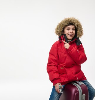 Hübscher europäischer junge, gekleidet in leuchtend rotem parka, der ein flugzeugspielzeugmodell hält und auf einen kopienraum mit weißem hintergrund zeigt, während er auf seinem gepäck sitzt. winterurlaub reisen, tourismuskonzept