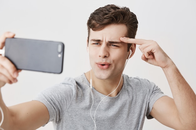 Hübscher europäischer Mann mit stilvollem Haarschnitt und blauen Augen, die weiße Kopfhörer tragen, Handy halten, für Selfie posieren, Gesichter machen, Hand hinter Augenbraue halten. Gesichtsausdrücke