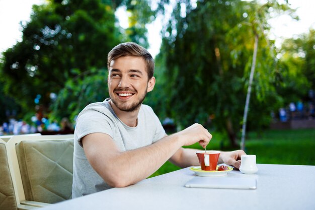 Hübscher fröhlicher lächelnder junger Mann, der am Tisch im Open-Air-Café mit Tasse Kaffee sitzt.