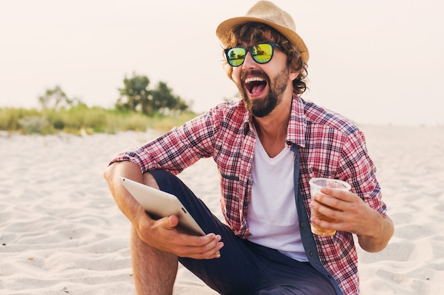 Hübscher fröhlicher Mann mit Bart im Strohhut, kariertem Hemd und stilvoller Sonnenbrille, die auf weißem Sand sitzt und Tablette verwendet