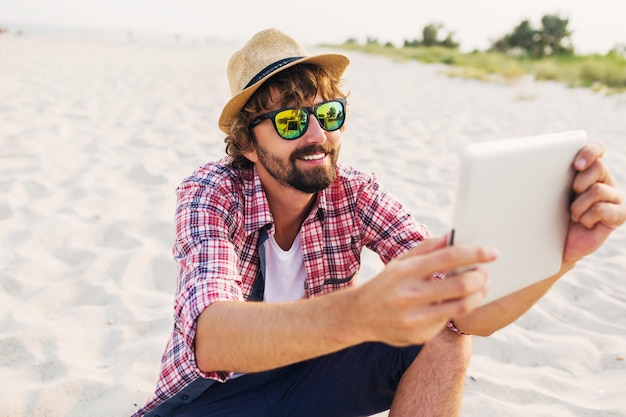 Hübscher fröhlicher Mann mit Bart im Strohhut, kariertem Hemd und stilvoller Sonnenbrille, die auf weißem Sand sitzt und Tablette verwendet