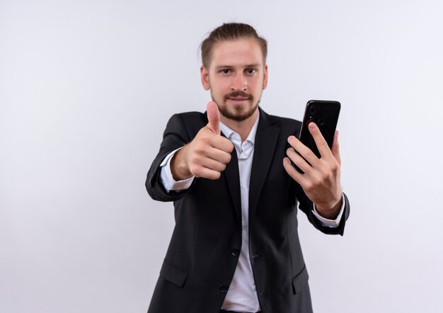 Hübscher Geschäftsmann, der Anzug hält, der lächelndes Smartphone hält, zeigt Daumen hoch, die über weißem Hintergrund stehen